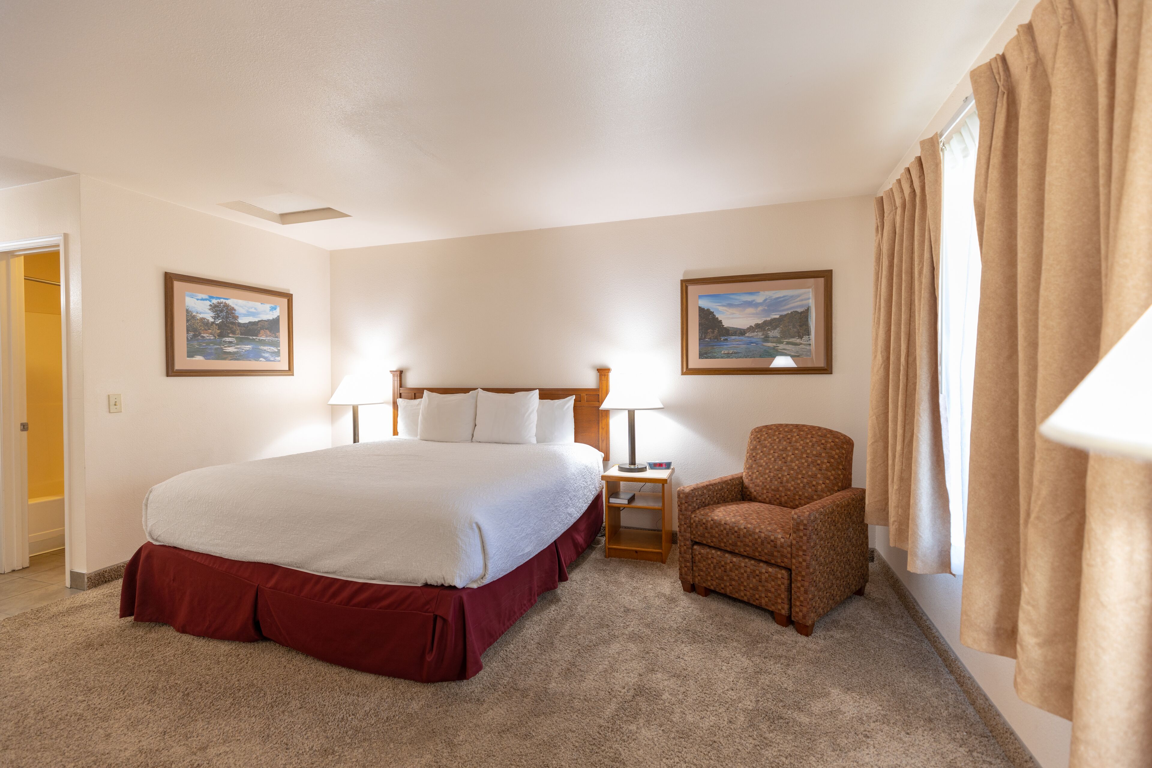 Habitación de hotel con una cama grande, una silla, dos cuadros enmarcados y una ventana con cortinas.