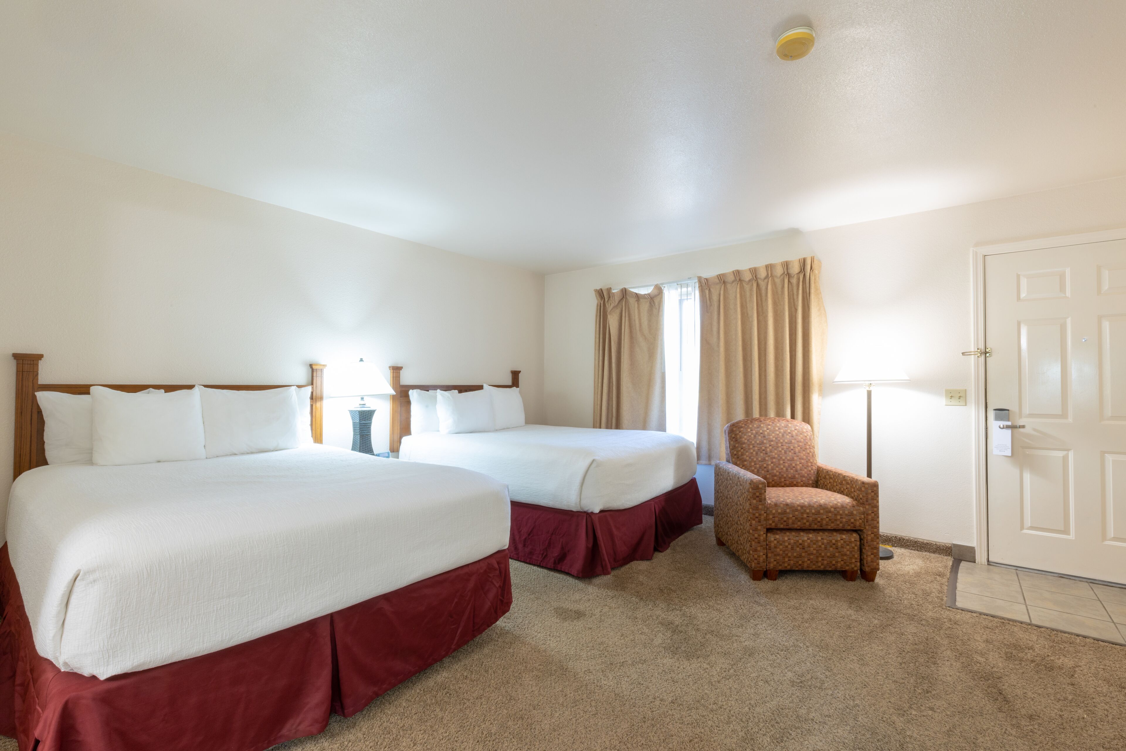 A hotel room with two beds, a chair, a lamp, and a door.