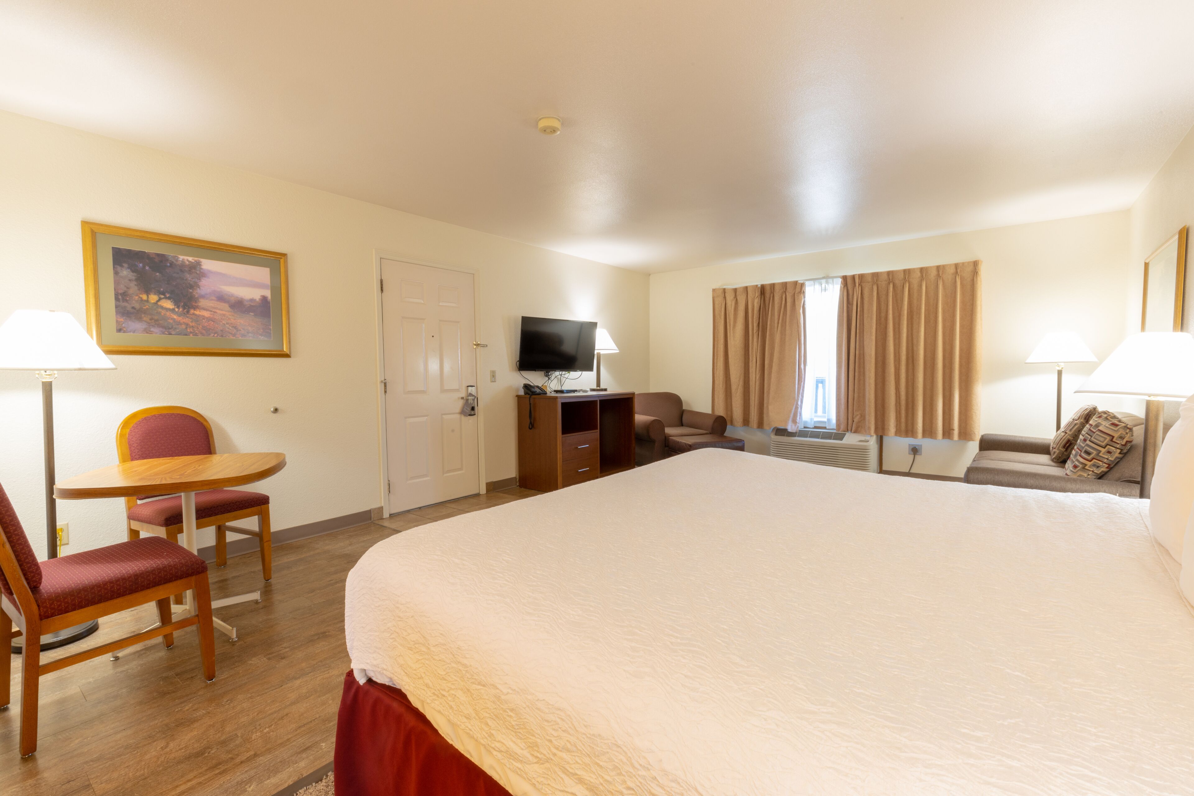 A hotel room with a large bed, a desk with a TV, a chair, a sofa, and a painting on the wall.