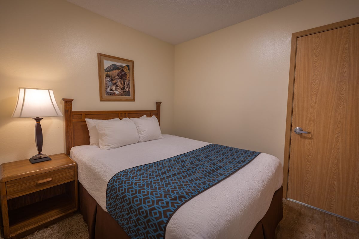 A hotel room with a bed, a nightstand with a lamp, a wooden door, and a framed picture on the wall.