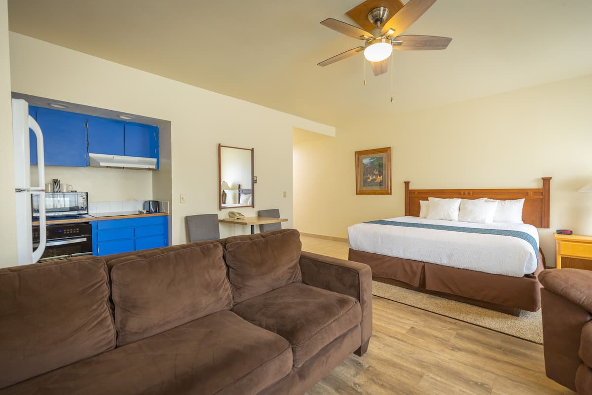 A compact hotel room with a kitchenette, a bed with a wooden headboard, a brown sofa, and a ceiling fan.