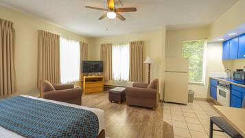 A hotel room with a bed, two armchairs, a TV, a small table, a ceiling fan, a kitchenette, and a window with curtains.