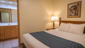 A hotel room with a bed, a nightstand with a lamp, a painting on the wall, and a bathroom with a sink and mirror.