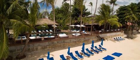 A beach resort with sun loungers, a pool, and palm trees.