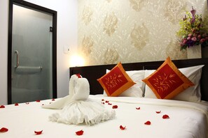 Habitación de hotel con cama, toalla en forma de cisne blanco, pétalos de rosa roja y almohadones decorativos.