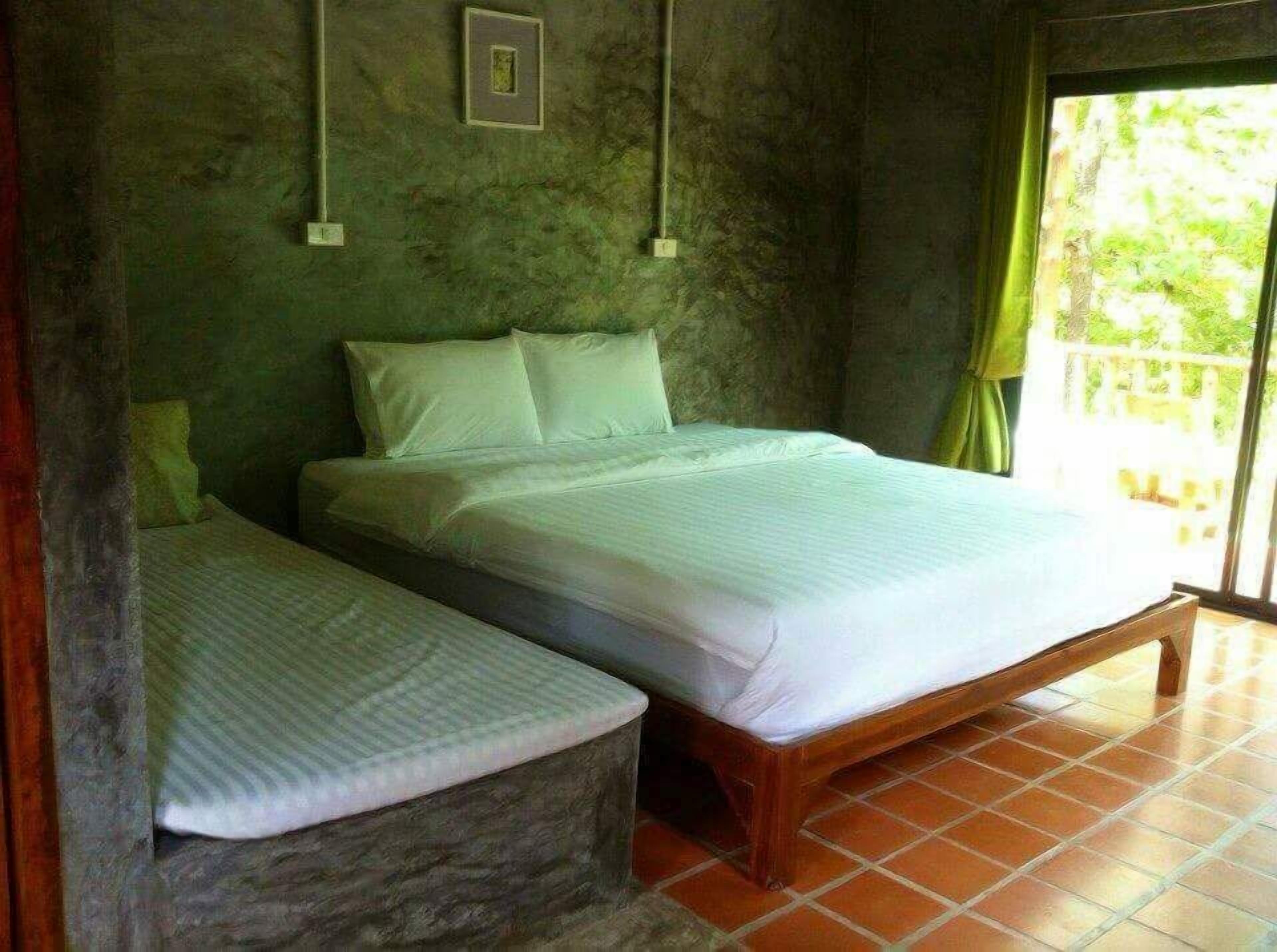 Un dormitorio con una cama individual, un banco, un cuadro pequeño en la pared y vistas a zonas verdes a través de una ventana.
