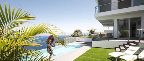 Un espace piscine extérieur moderne offrant une vue dégagée sur l’océan, un bâtiment à deux étages doté de balcons et une pelouse artificielle.