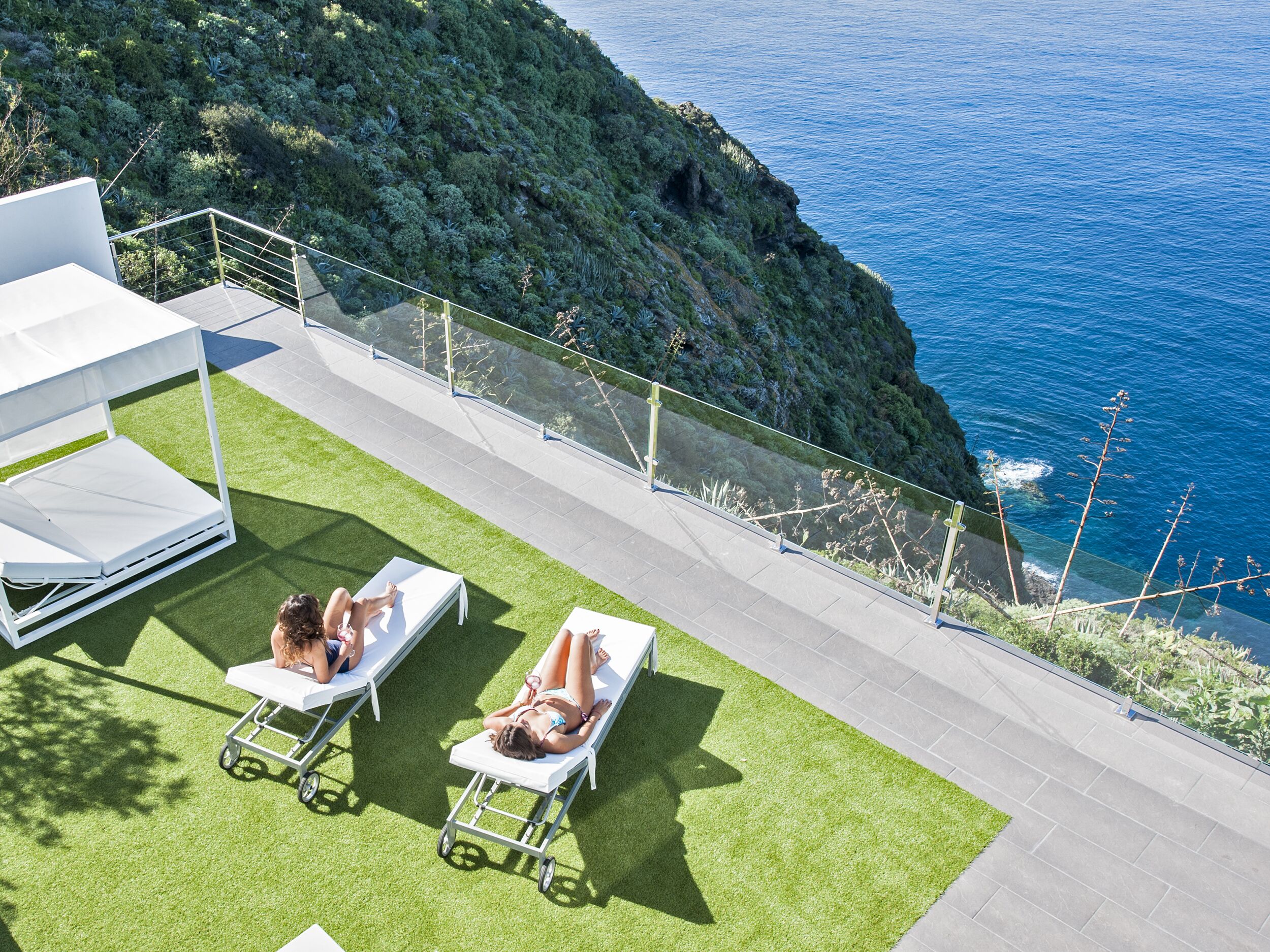 Una terraza en la azotea con dos tumbonas, un sofá blanco para exteriores y una barandilla de cristal con vistas a un acantilado costero y al océano.