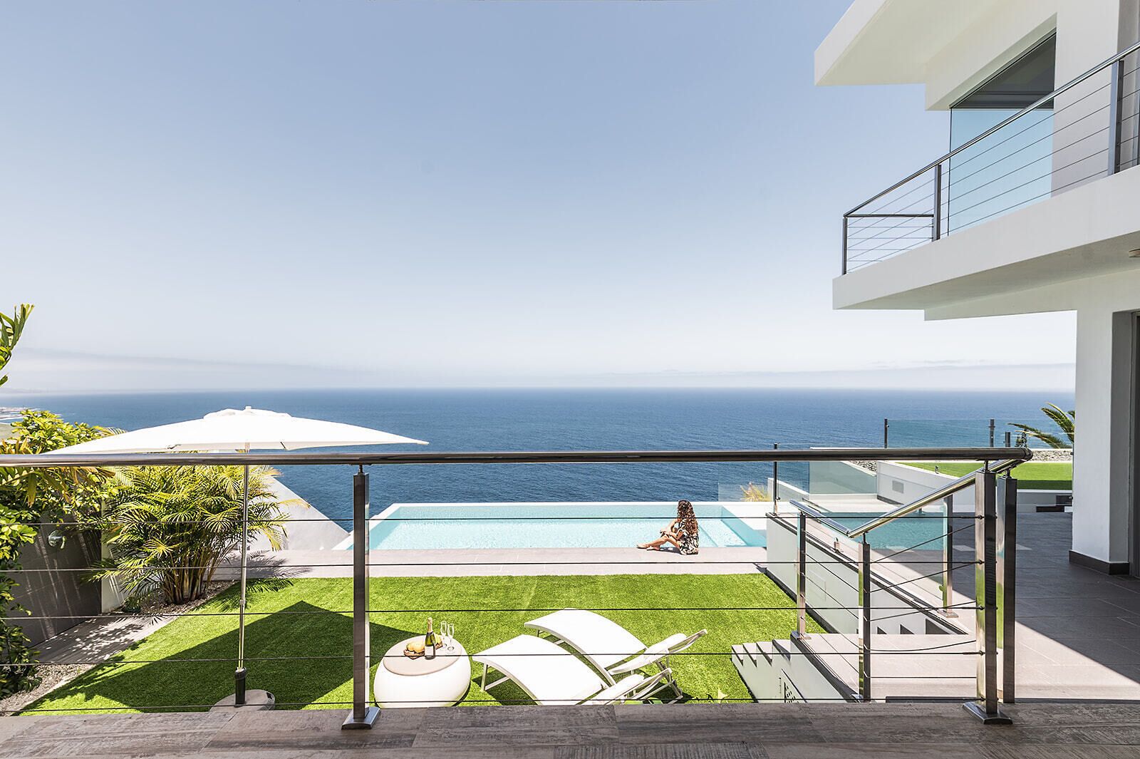 A modern outdoor pool area with a clear view of the ocean, a grassy lawn, and a white umbrella.