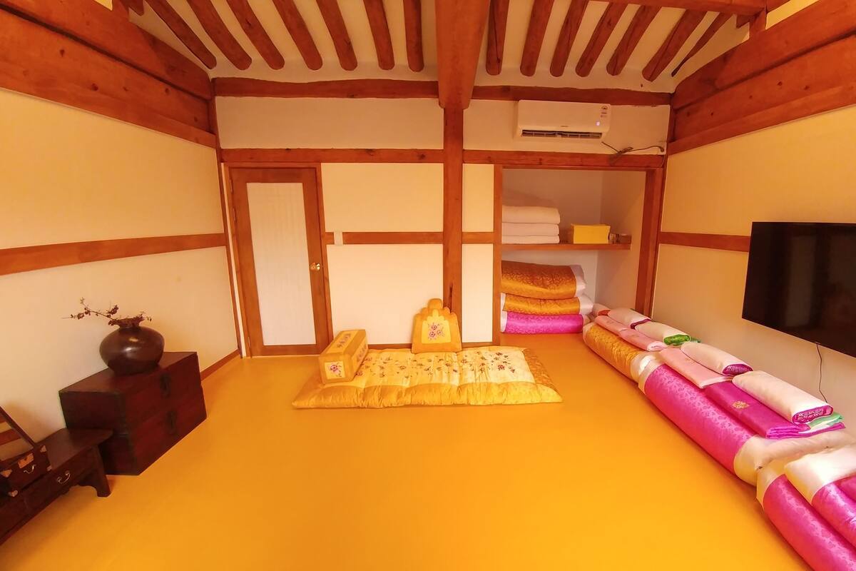 A traditional Japanese-style room with tatami flooring, a futon bed, a television, and a wooden shelf with folded textiles.