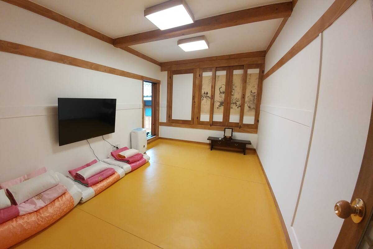 A room with a flat-screen TV mounted on the wall, a wooden bench, and a futon bed with pillows.