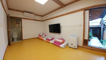 Una habitación con piso amarillo, un inodoro, una cama con ropa de cama rosa y blanca, un purificador de aire Samsung y un televisor en la pared.