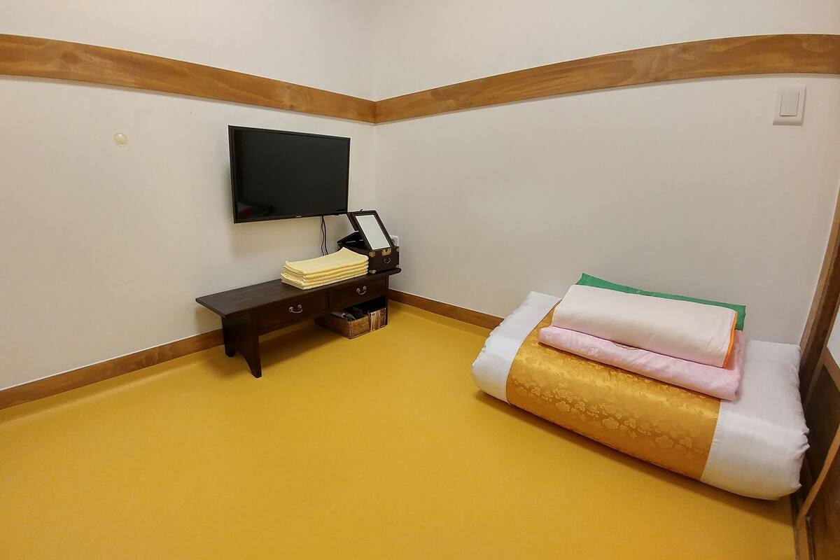 A room with a yellow floor, a wooden bench, a small table with a TV, and a bed with pillows.