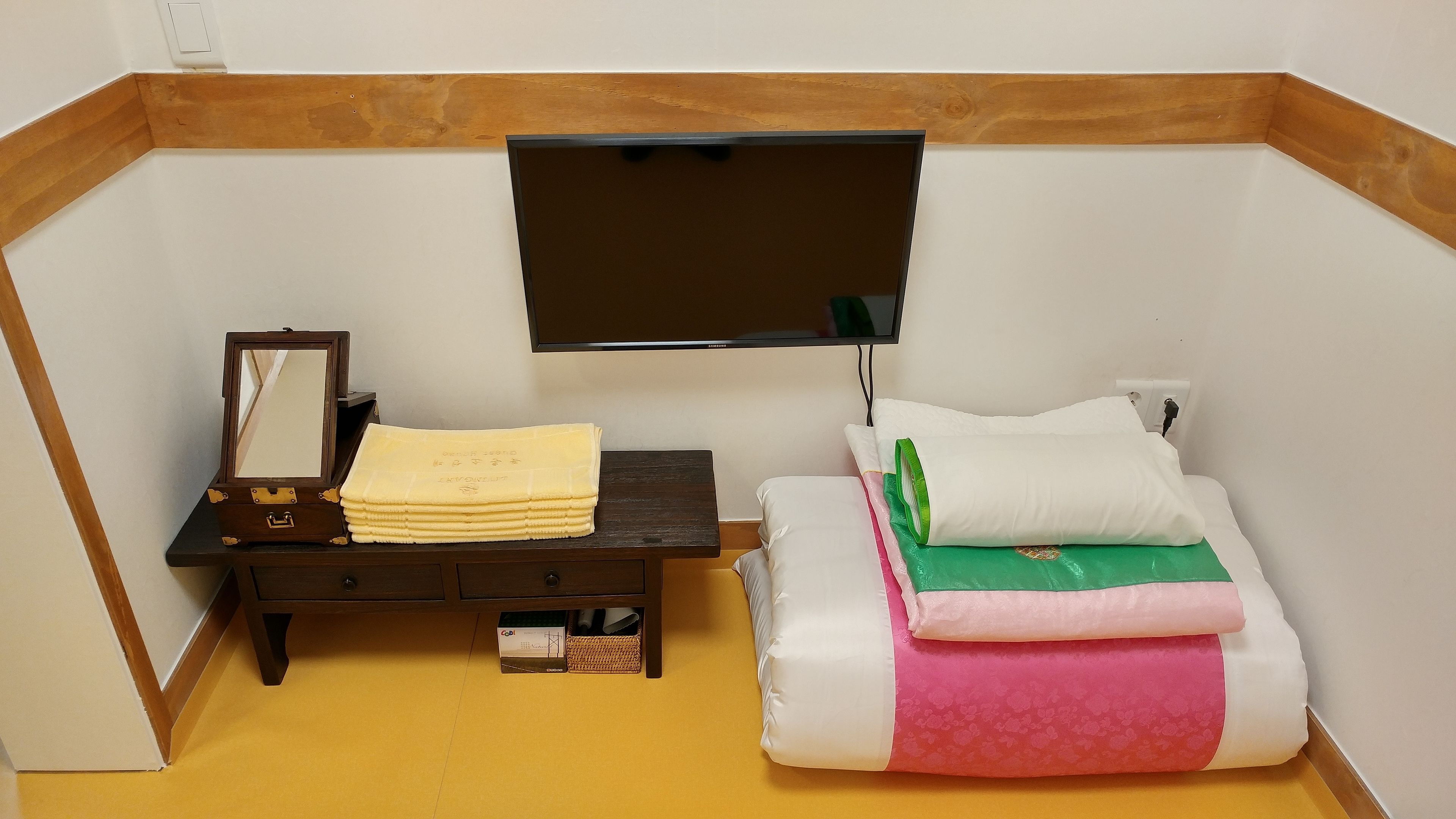 A small, compact room with a bed, a bedside table with a TV, a mirror, and a stack of towels.