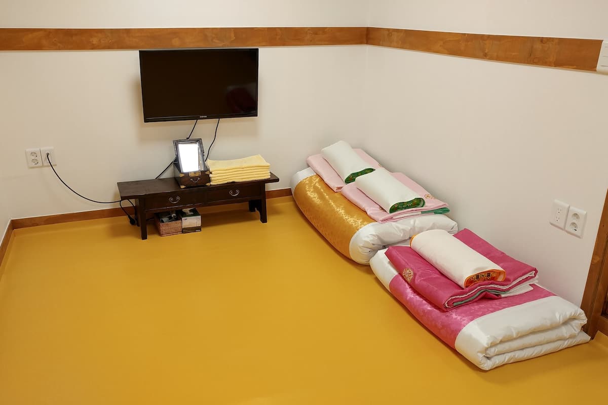 A room with a flat-screen TV mounted on the wall, a small wooden table with a lamp, and a bed with rolled-up towels.