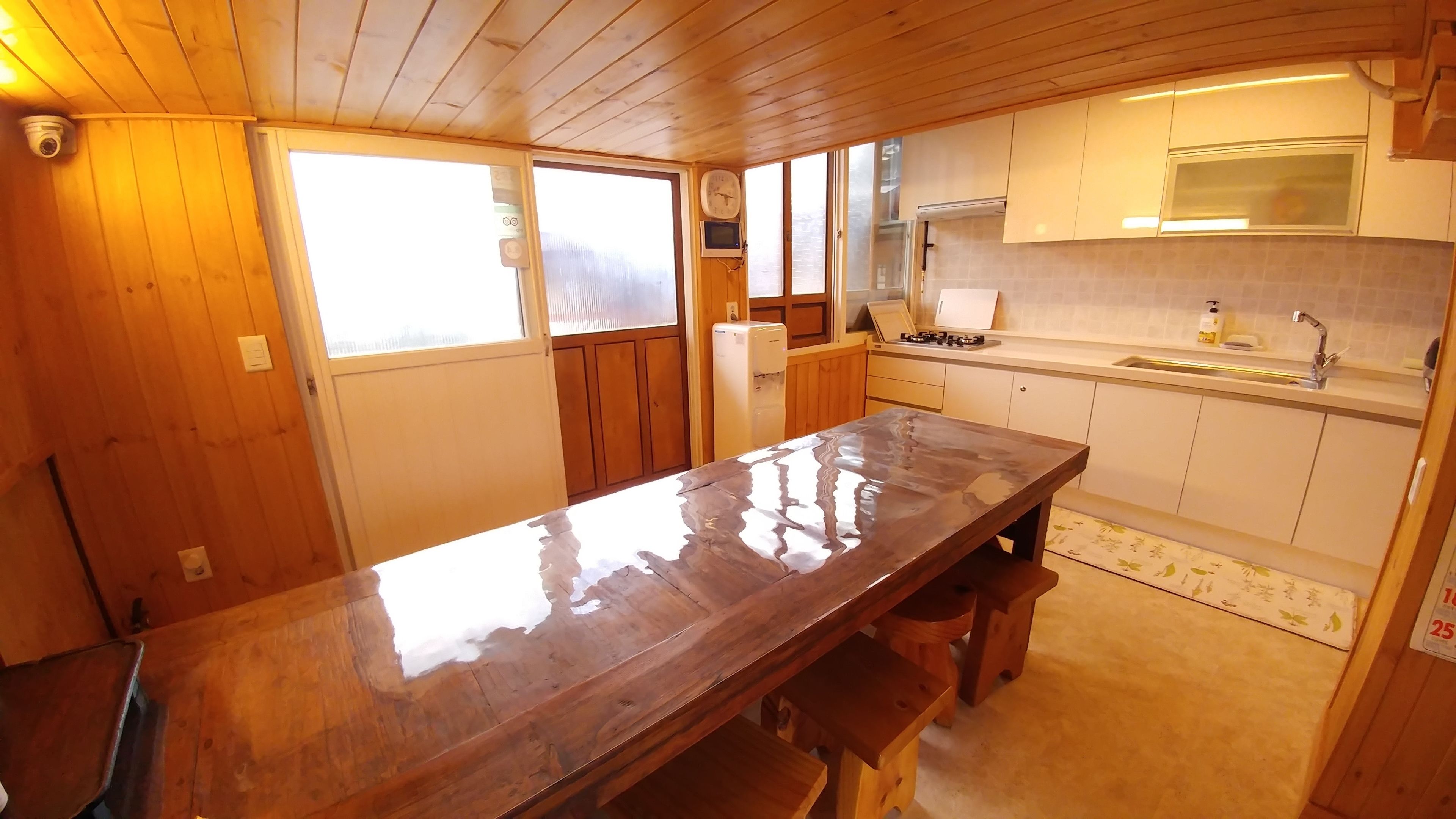 Un interior de madera con una larga mesa, una zona de cocina con fregadero y hornilla, y un reloj en la pared.