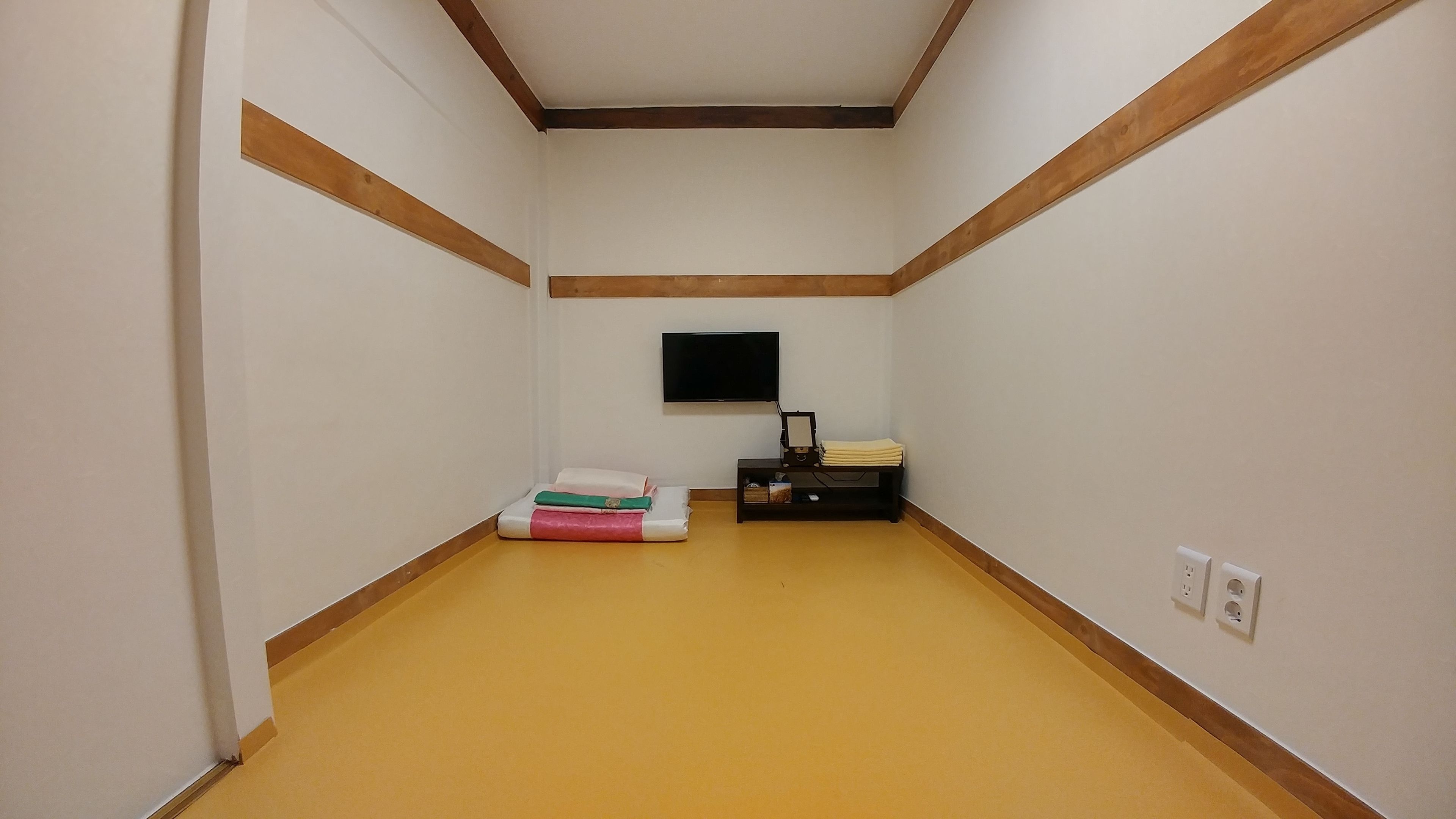 A narrow hallway with a yellow floor, white walls, and wooden trim. A flat-screen TV is mounted on the wall, and there is a small table with folded towels and a box.
