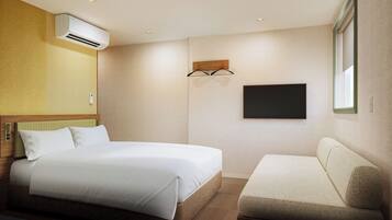 A hotel room with a bed, a sofa, an air conditioning unit, a wall-mounted TV, and a window.