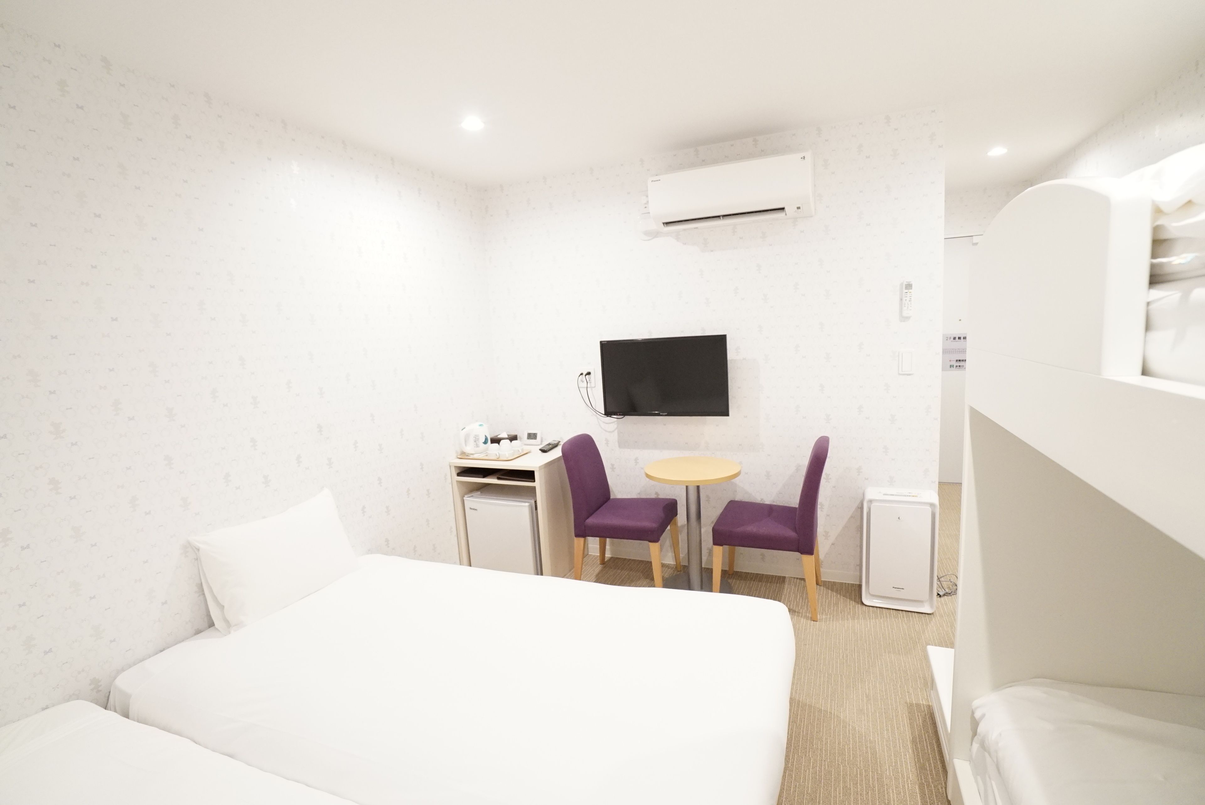 A hotel room with a bed, a desk with a computer, a TV, and two purple chairs.