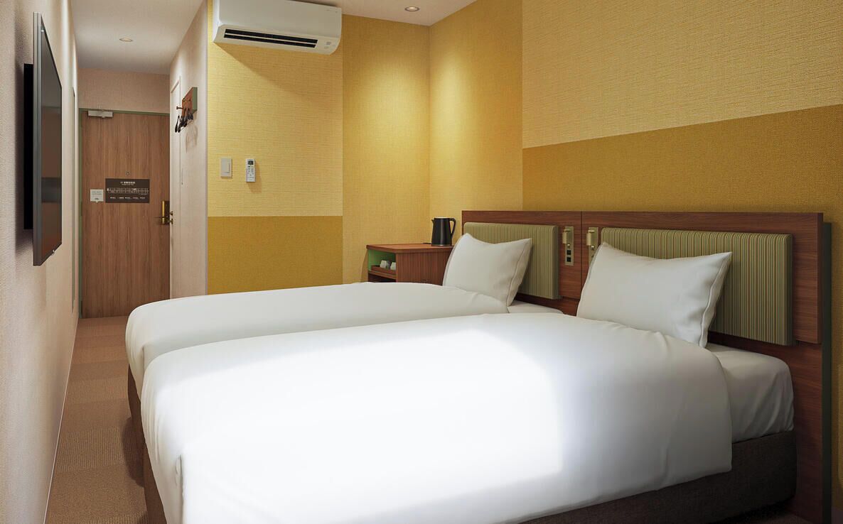 A hotel room with two beds, a wooden door, an air conditioning unit, a nightstand, and a small table with a lamp.
