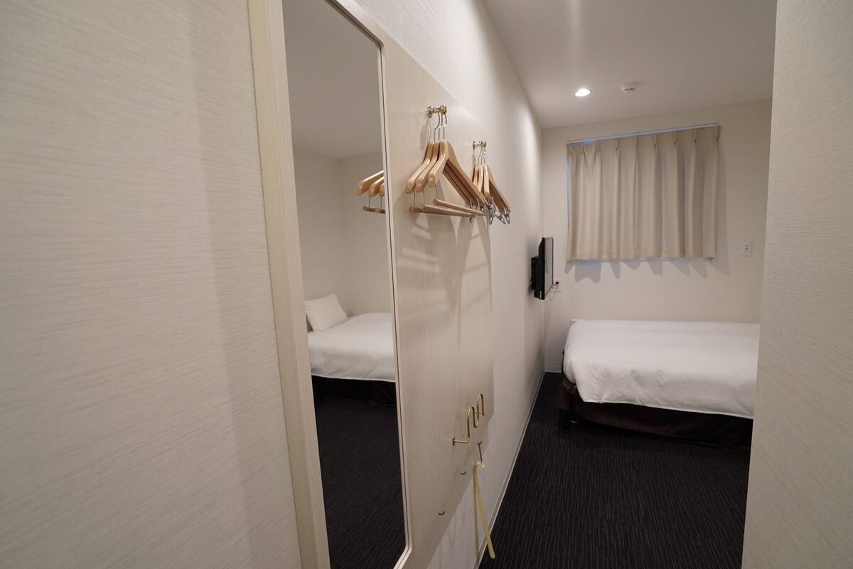 Hotellihuone, jossa on kaksi sänkyä, vaatekaappi puisten henkareiden kanssa, taulu-tv ja ikkuna, jossa on verhot.