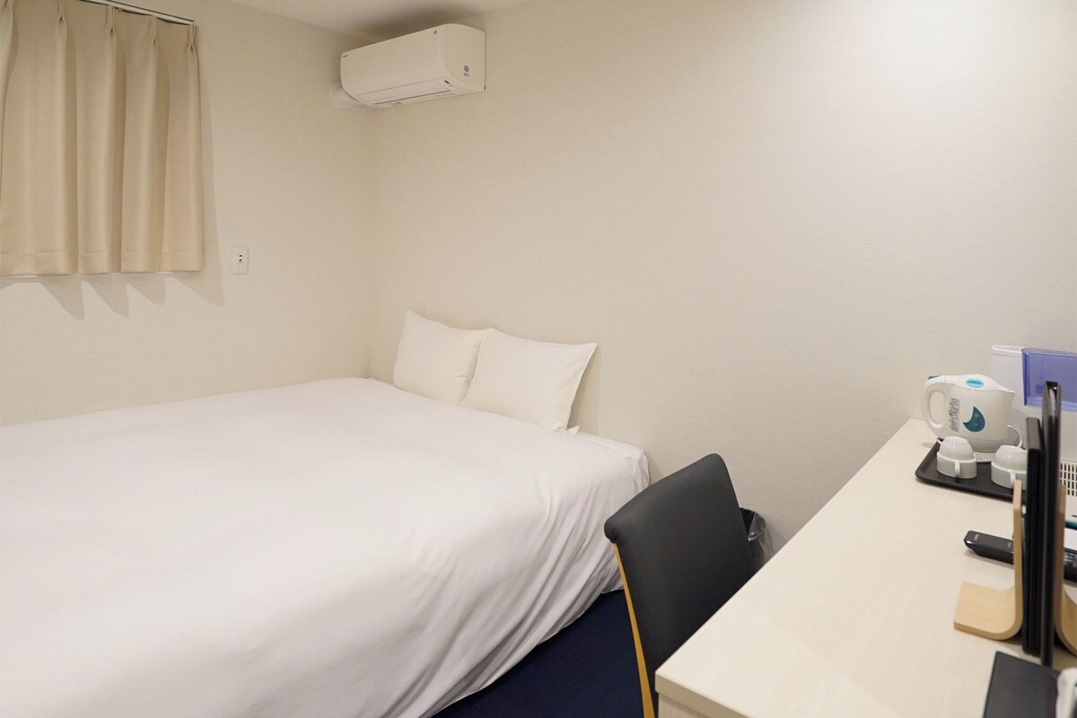 Quarto de hotel com cama, escrivaninha com cafeteira e uma cadeira.