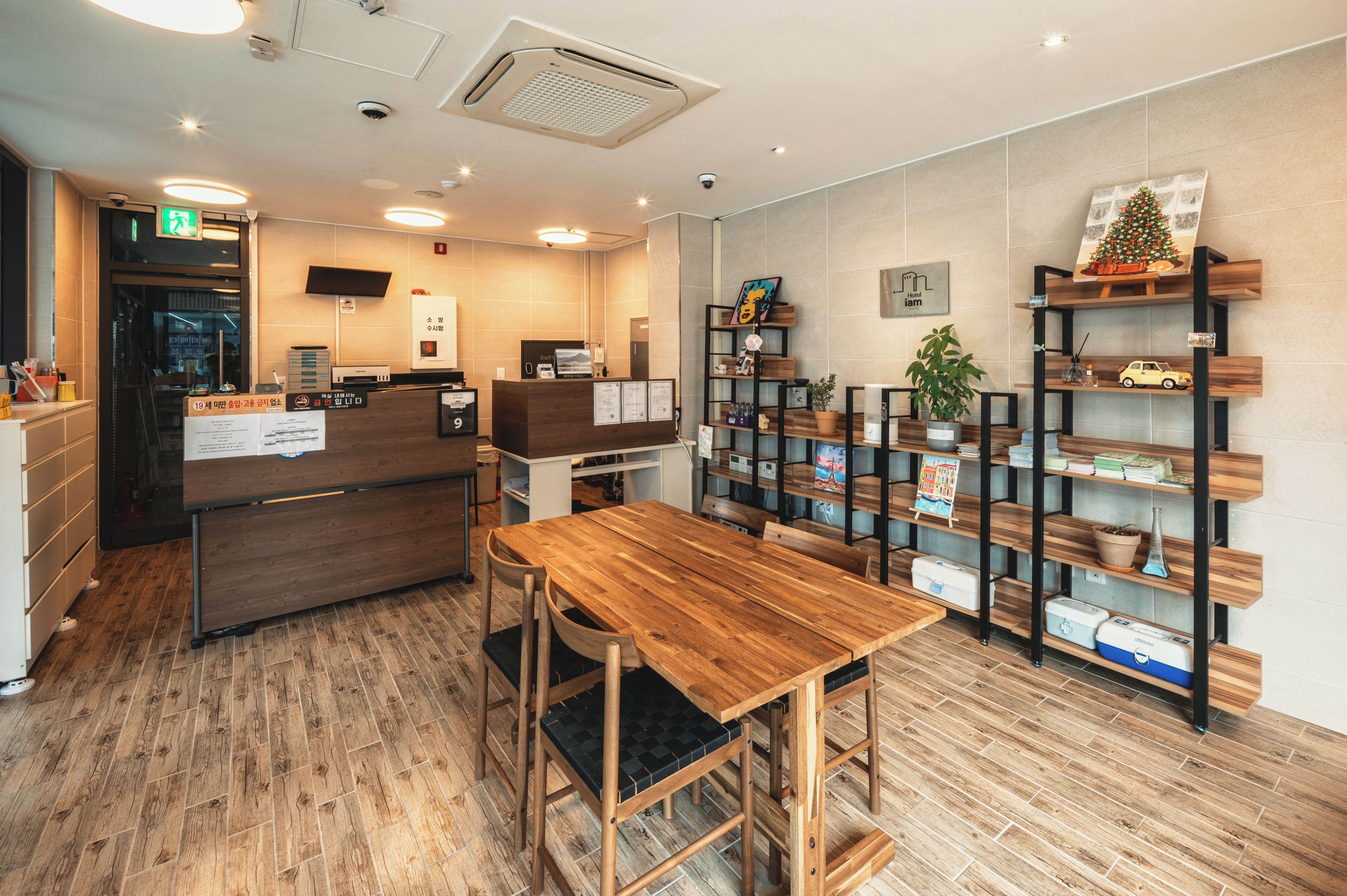 Un área de recepción con un mostrador de madera, un espacio de espera con una mesa y sillas de madera, y una estantería con diversos objetos.