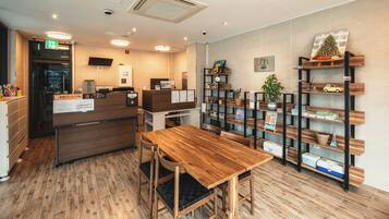 Un área de recepción con un mostrador de madera, un espacio de espera con una mesa y sillas de madera, y una estantería con diversos objetos.