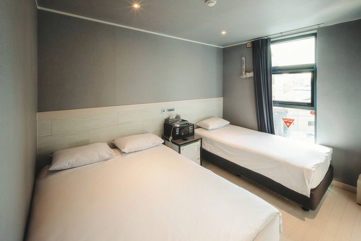 A hotel room with two beds, a small bedside table with a radio, and a window with curtains.