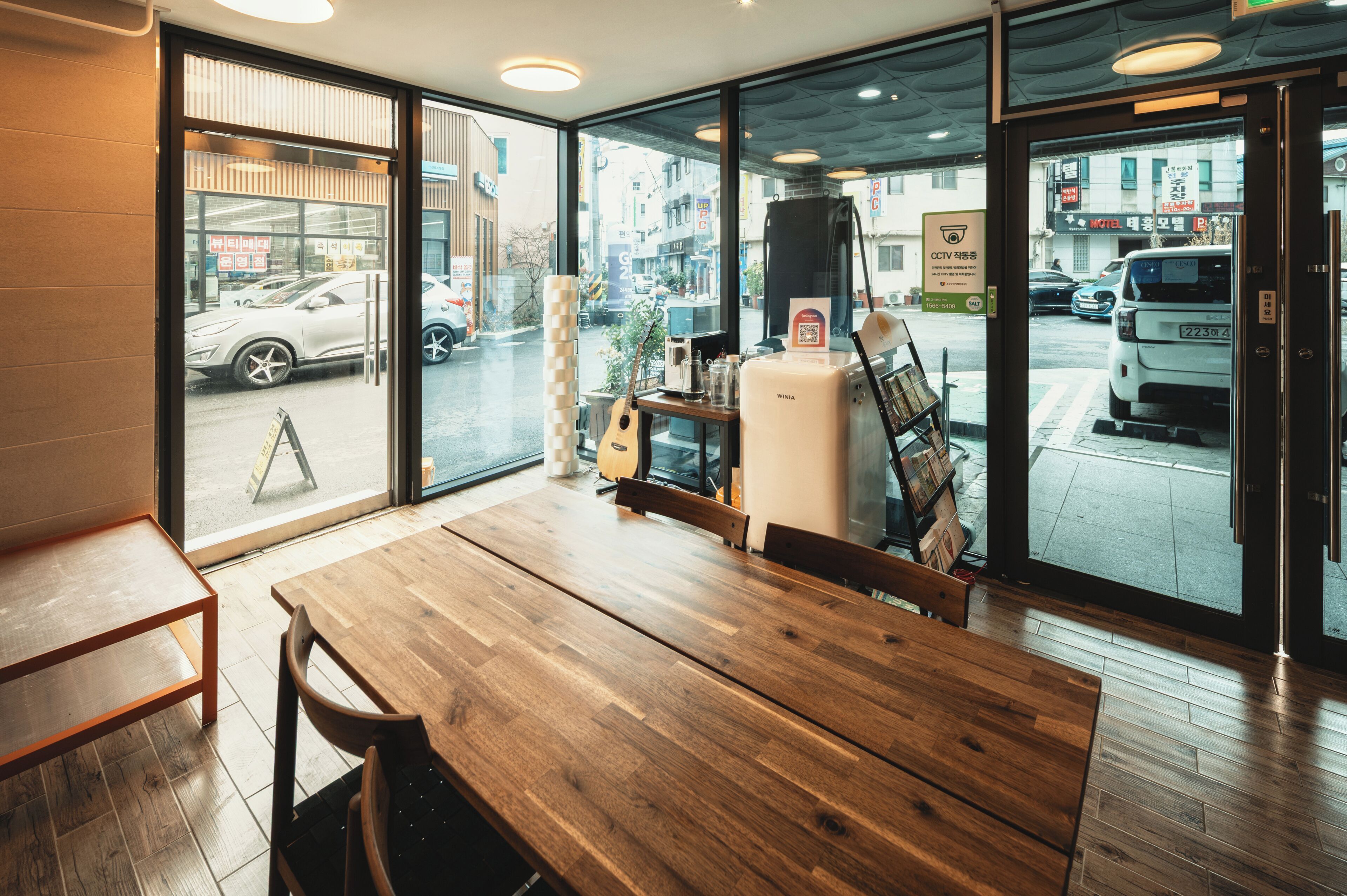 Une table et des chaises en bois dans un café aux grandes fenêtres, une guitare et un appareil à café.