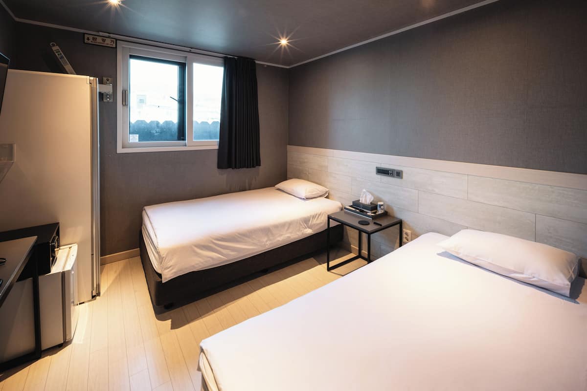 A hotel room with two beds, a small table, a refrigerator, and a window with curtains.