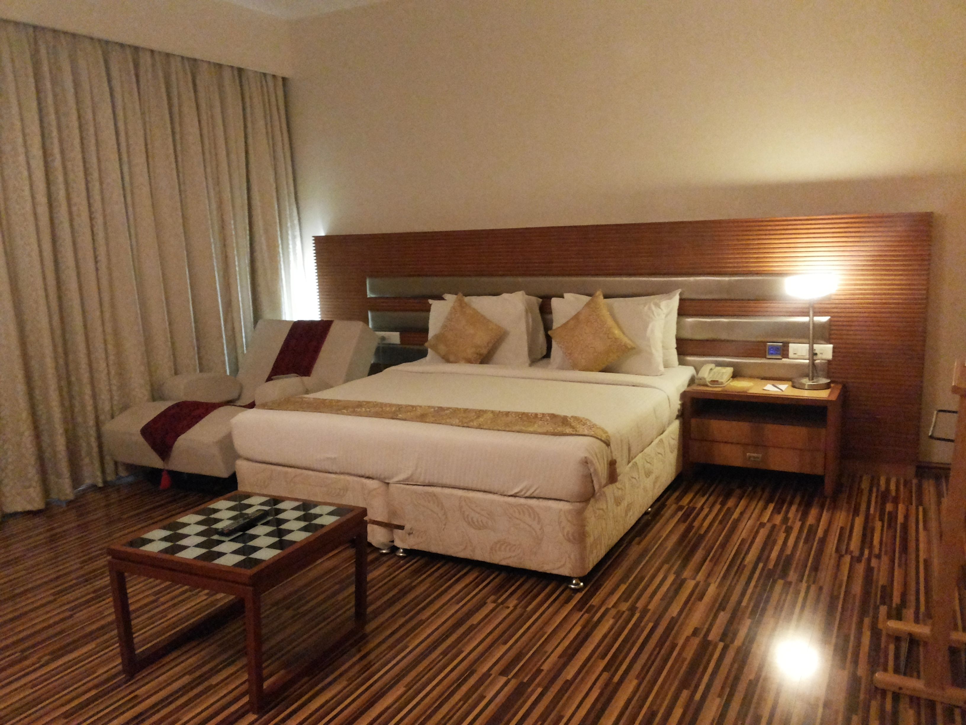 A hotel room with a large bed, a bedside table with a lamp, a small table with a chessboard, and a wooden floor.
