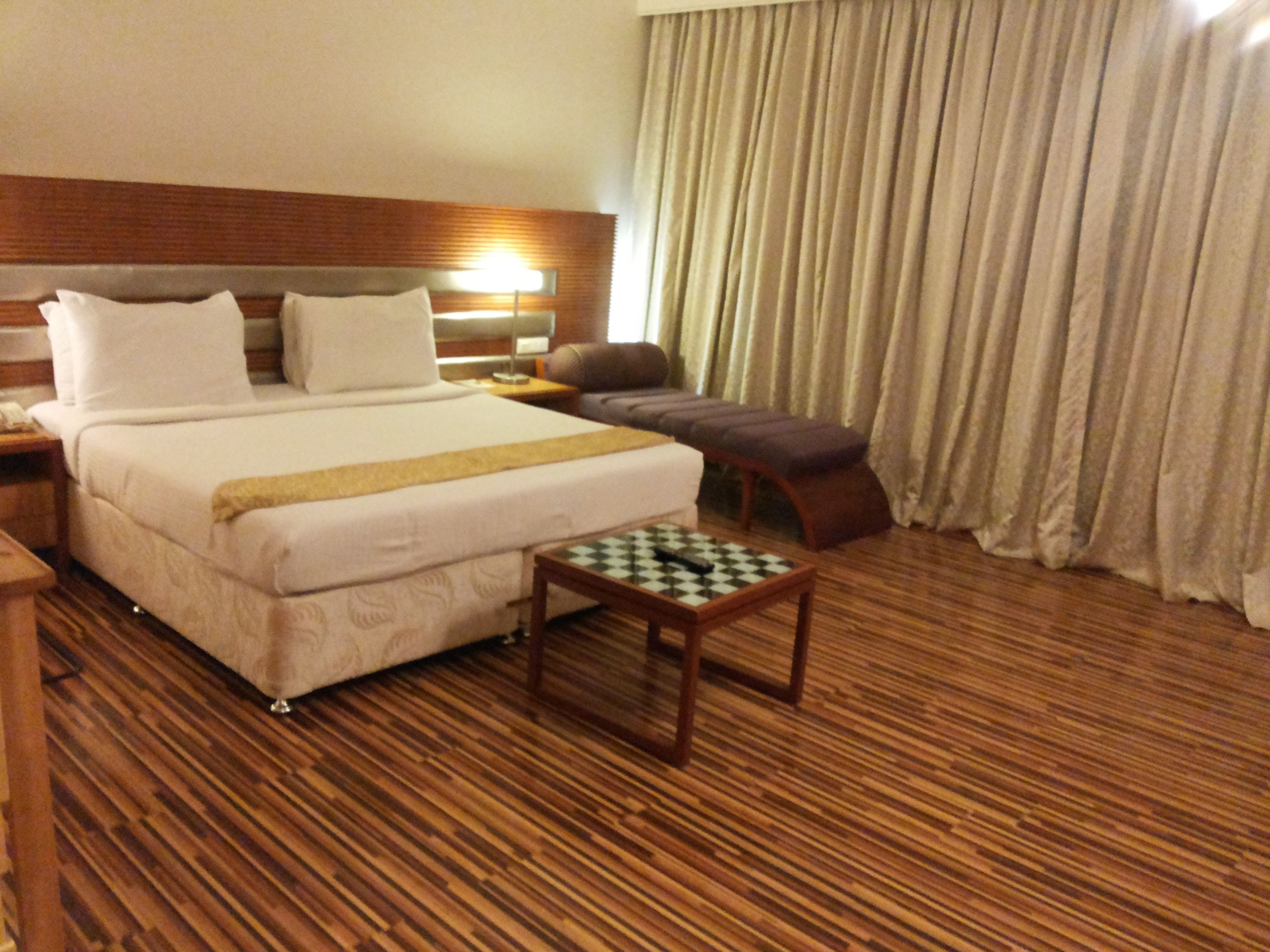 A hotel room with a large bed, a wooden floor, a small table, a chair, and a window with curtains.