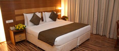 A hotel room with a large bed, wooden headboard, a nightstand with a lamp, and a window with curtains.