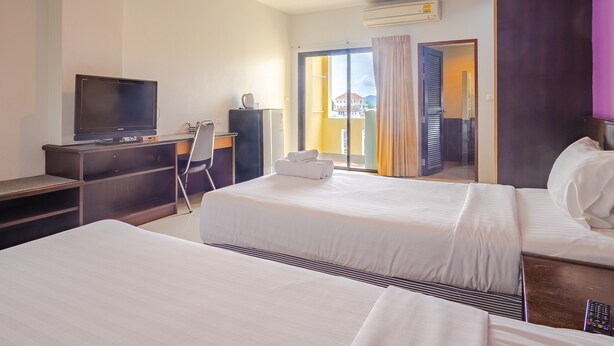 A hotel room with two beds, a desk with a television, a chair, and a view of a building through a glass door. - AJ Residence (Ratsada)