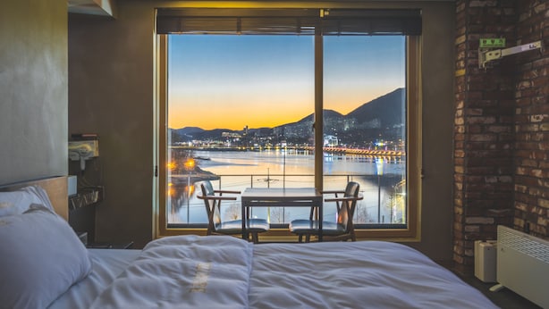 A hotel room with a large window offering a view of a cityscape and a body of water at sunset. There is a bed with white linens and a small table with two chairs. - B&F Hotel (Yeosu)