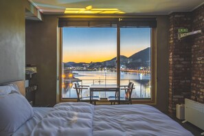A hotel room with a large window offering a view of a cityscape and a body of water at sunset. There is a bed with white linens and a small table with two chairs. - B&F Hotel (Yeosu)