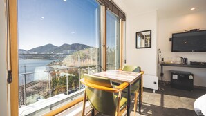 A hotel room with a large window offering a view of a body of water and hills. There is a dining table with chairs, a flat-screen TV, and a small kitchenette area. - B&F Hotel (Yeosu)