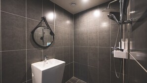 A modern bathroom with dark tiles, a white sink, and a shower with a handheld showerhead. - B&F Hotel (Yeosu)