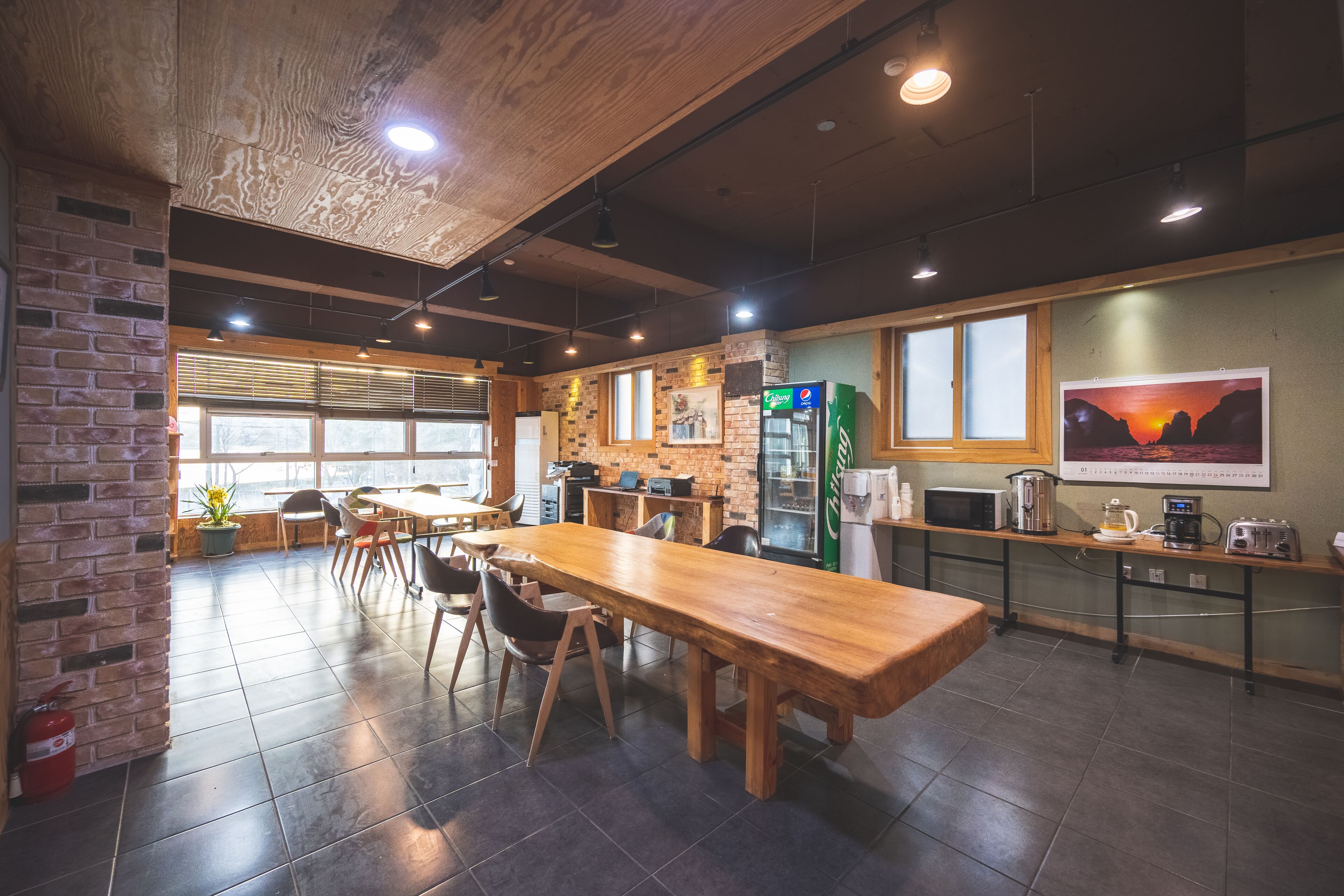 A spacious cafe with a long wooden table, brick walls, and large windows.