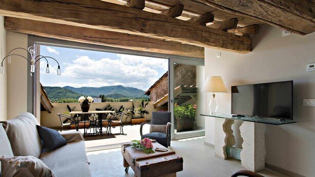 A spacious living area with a sofa, a coffee table, and a TV mounted on the wall. There is a balcony with a table and chairs, and a view of the mountains. - Argenter, deluxe penthouse with terrace (Olot)