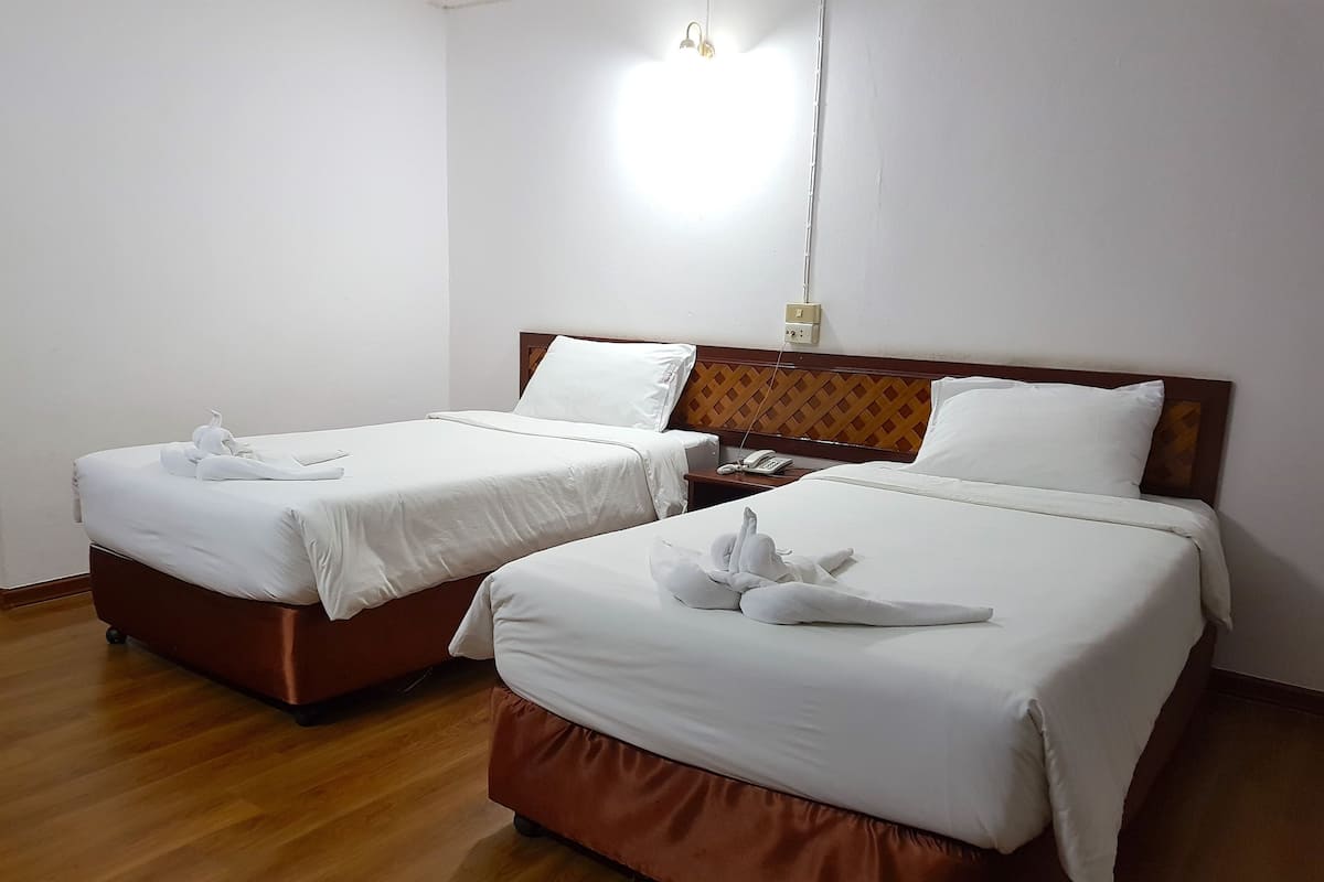 Ein Hotelzimmer mit zwei Betten, jeweils mit weißen Laken und Handtüchern. Es gibt ein hölzernes Kopfende mit einem gemusterten Design, einen Nachttisch mit einem Telefon und eine an der Wand befestigte Leuchte.
