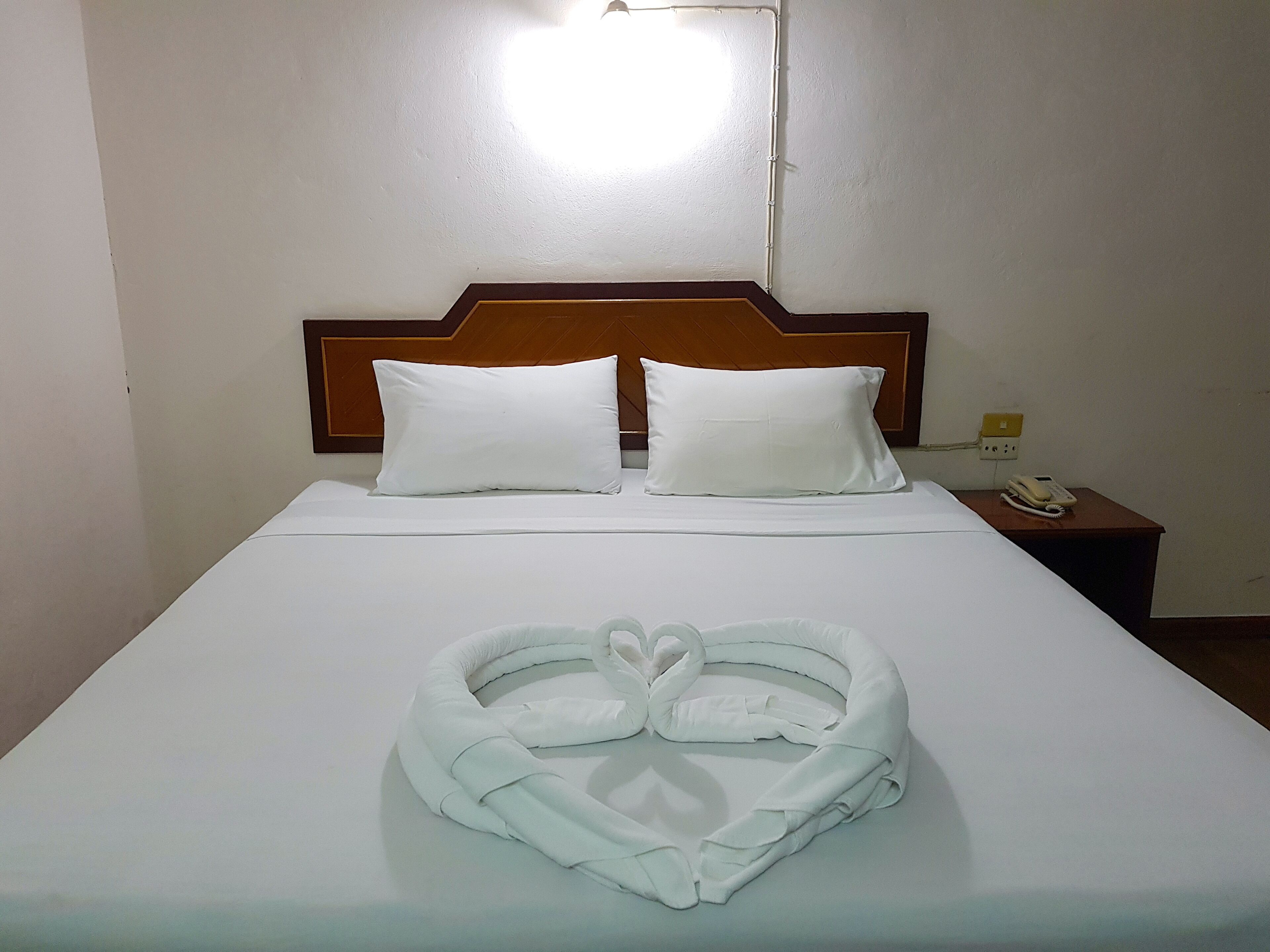 A neatly made bed with white linens and pillows, a heart-shaped towel arrangement, and a bedside telephone on a wooden nightstand.