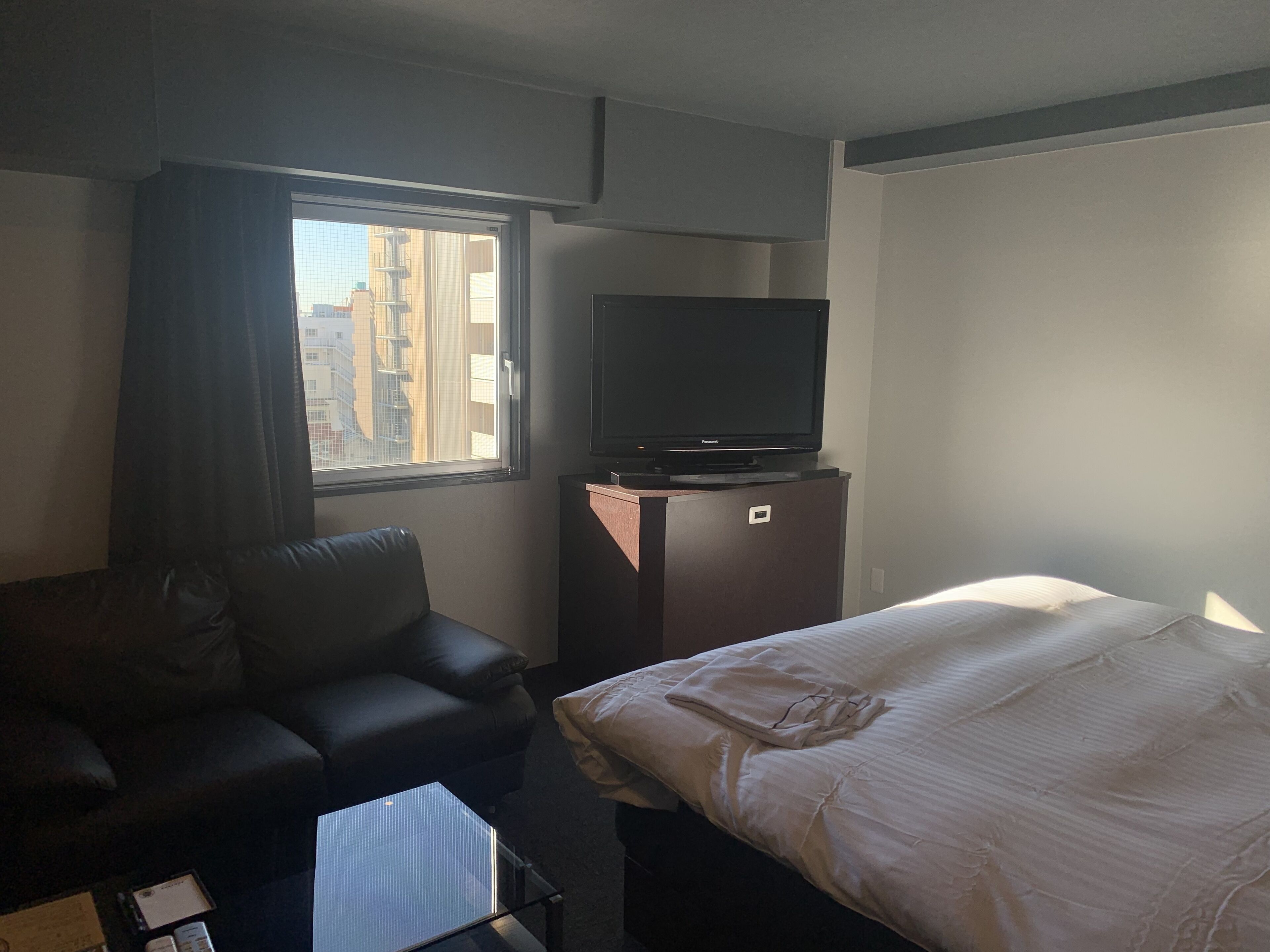 A hotel room with a bed, a TV on a cabinet, a couch, and a window with a view of buildings.