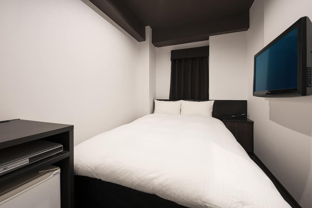 A hotel room with a bed, a flat-screen TV mounted on the wall, a small desk with a computer, and a heater.