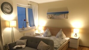 A bedroom with a bed, a bedside table with a lamp, a window with curtains, a wall clock, and a painting of a beach scene.