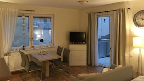 A compact living space with a dining table, chairs, a sofa, a TV, and a balcony with curtains.