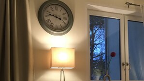 A living room with a sofa, a wall clock, a floor lamp, a window with a view of trees, and a vase with a flower.