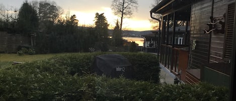 En trädgård med en överbyggd grillplats, tät buskvegetation och utsikt över en sjö vid solnedgången.