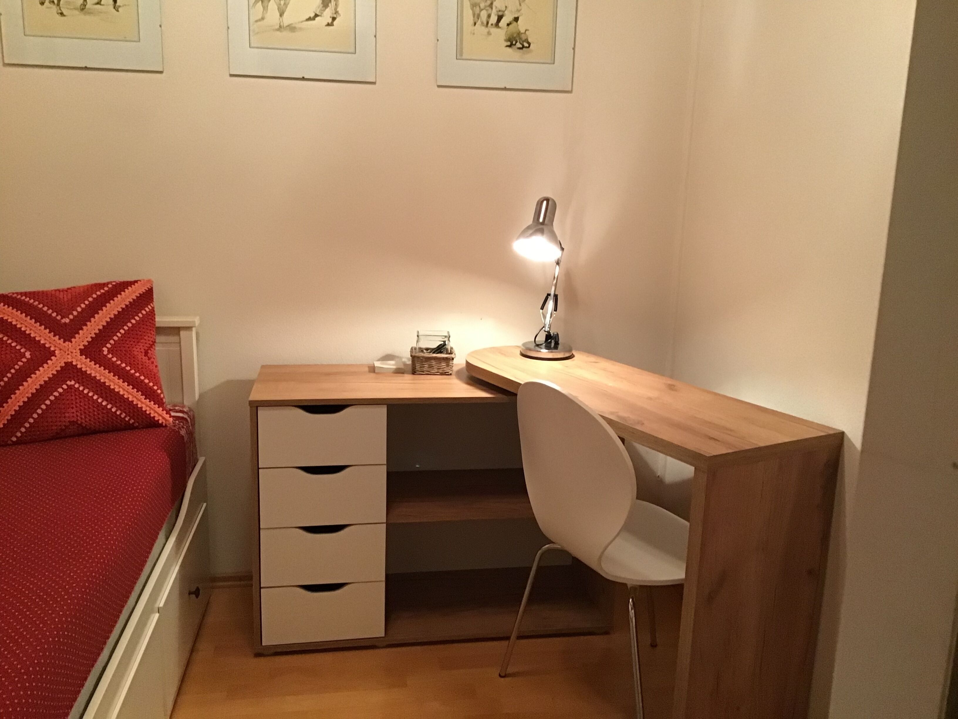 Une chambre avec un bureau en bois, une chaise blanche, un coussin rouge, une lampe et un petit panier.